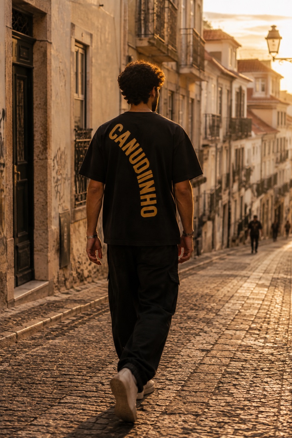 Pessoa de costas a descer uma rua de Lisboa ao fim da tarde, com a palavra Canudinho bordada a dourado nas costas da t-shirt preta